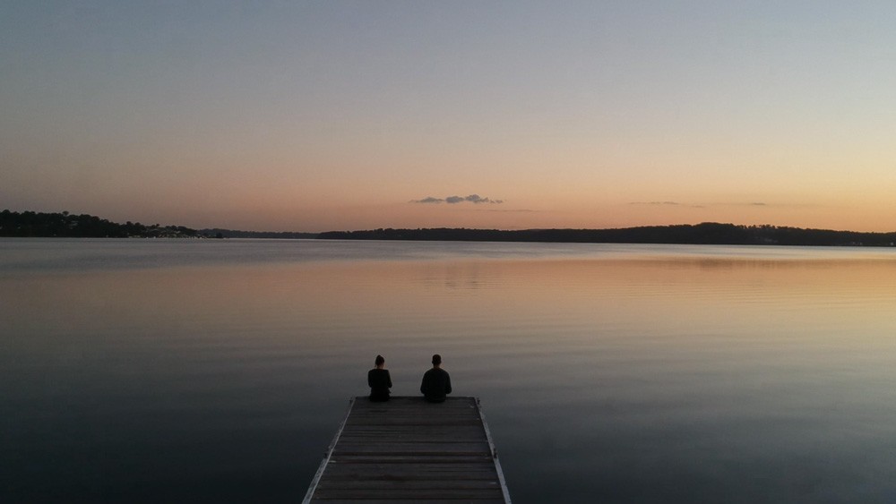 dos personas sentadas y de espaldas en un puente y delante paisaje de una puesta de sol en un lago